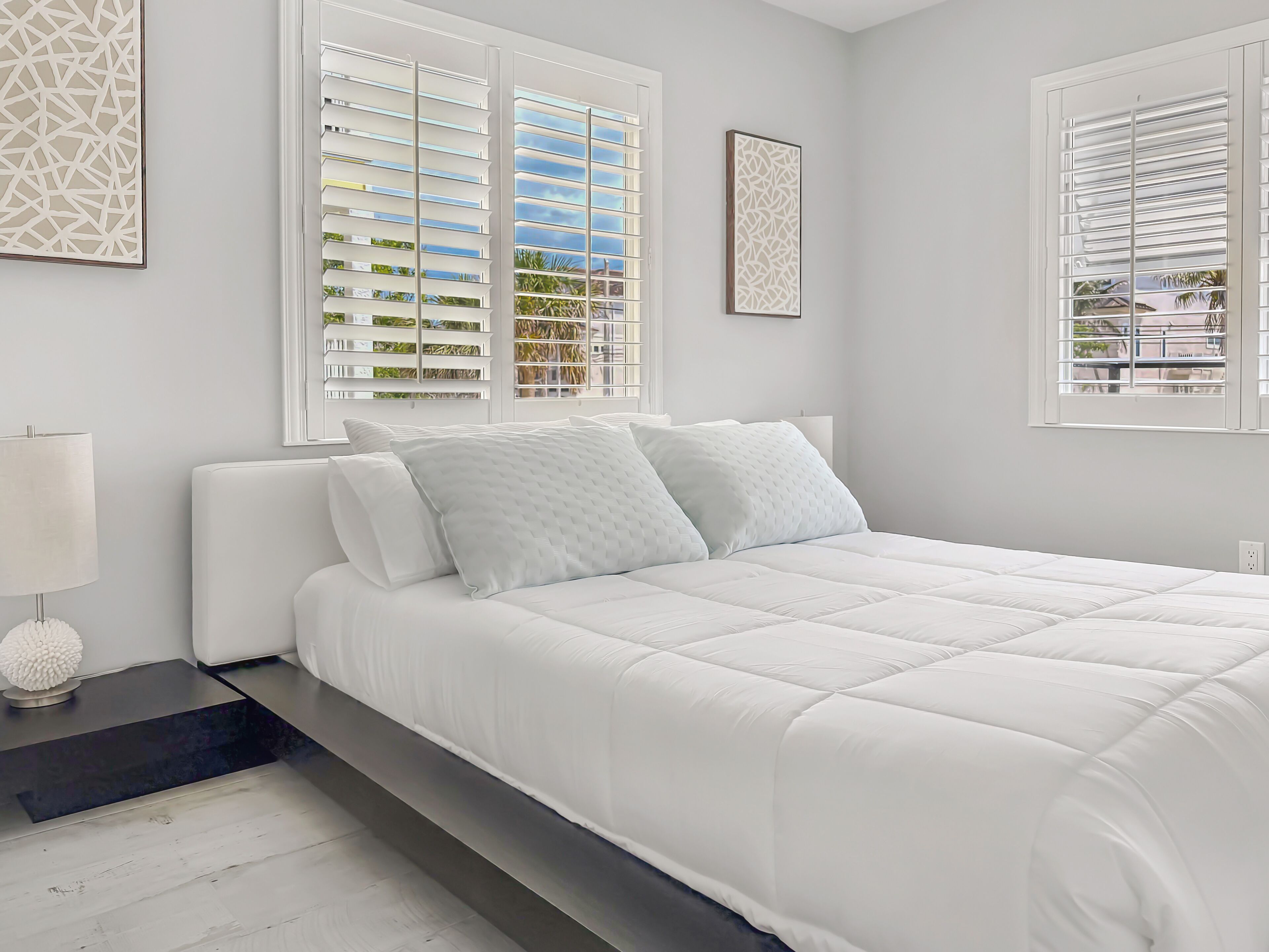 Una cama bien hecha con ropa blanca, una mesita de noche con lámpara y una ventana con persianas.