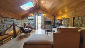 Ein gemütliches Dachzimmer mit Steinwand, Holzbalkendecke und einem großen Fenster. Es gibt Ledersofas und einen kleinen Tisch.