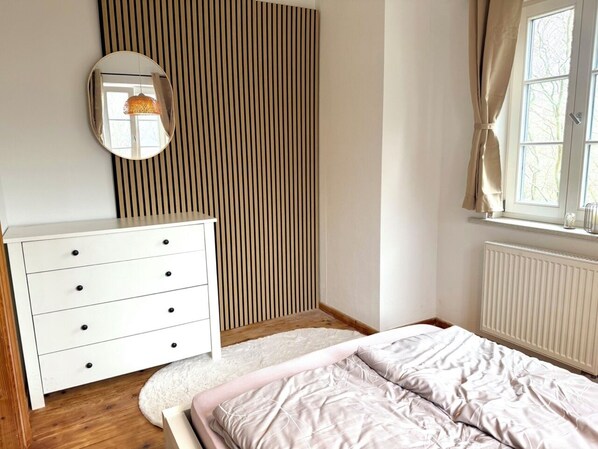 Une chambre à coucher avec un lit, une commode, un miroir, un radiateur et une fenêtre avec des rideaux.