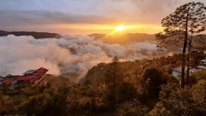 A mountainous landscape with a sunrise, clouds below the treetops, and buildings in the foreground.