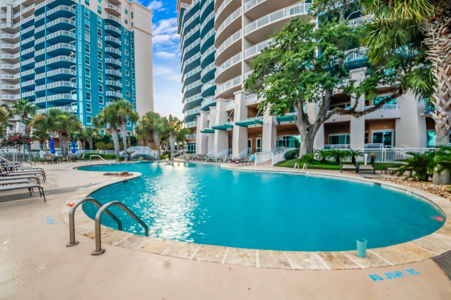 A swimming pool surrounded by multi-story buildings with balconies, palm trees, and a clear sky.