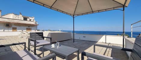 An outdoor terrace with a table and chairs, a large umbrella, and a view of the sea.