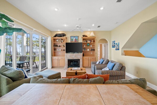 A spacious living room with a fireplace, a television, and a large window overlooking a deck. - Banana Bay-Bayfront, Private Pool, East End of SGI (Eastpoint)