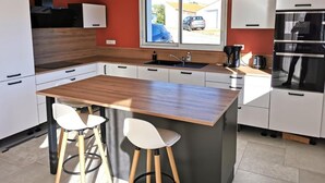 Une cuisine moderne avec un îlot central en bois, des armoires blanches et une grande fenêtre donnant sur une voiture et une maison.