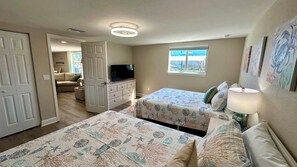 A bedroom with two beds, a television, a window, and a door leading to another room.