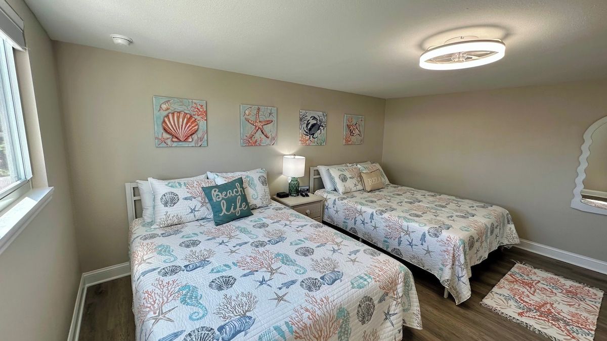 A bedroom with two beds, each with a beach-themed bedspread, a bedside table with a lamp, and wall art depicting sea creatures.