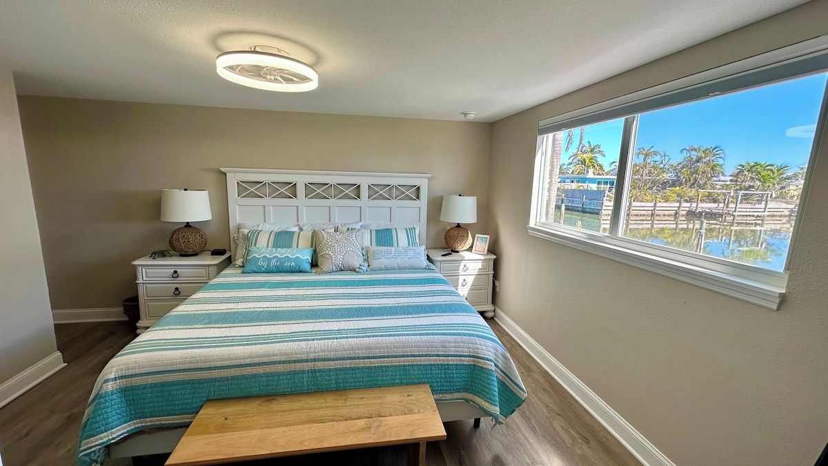 Une chambre à coucher comprenant un lit, des tables de chevet, des lampes, un banc et une fenêtre donnant sur des palmiers.