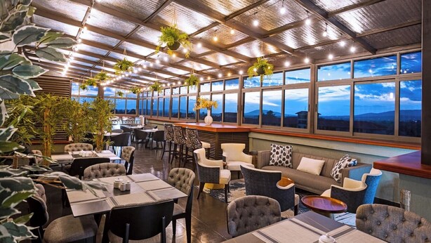 A restaurant with a high ceiling, hanging plants, and large windows offering a view of the mountains. - Clayton Luxury Suite (Machakos)