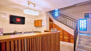A hotel reception area with a wooden reception desk, a mounted television displaying the CNN logo, a staircase with a metal railing, and a 'Welcome' sign. - Clayton Luxury Suite (Machakos)