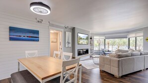 Un salón moderno con una mesa de comedor de madera, sillas blancas, un sofá beige y un ventanal con vistas a palmeras.