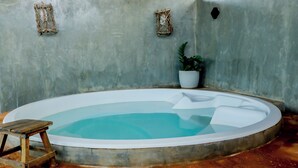 A small, round hot tub with a wooden stool, a potted plant, and two wall-mounted decorative items in a room with a concrete wall and brick floor.