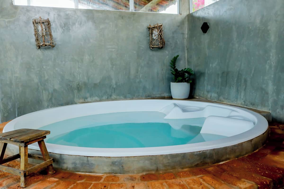 Un hidromasaje pequeño y redondo con un taburete de madera, una planta en maceta y dos elementos decorativos en la pared, en una habitación con pared de concreto y suelo de ladrillo.