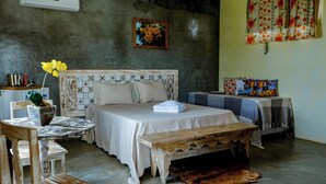 A bedroom with a bed, a wooden bench, a table with a tablecloth, a chair, a potted plant, and a wall-mounted air conditioner.