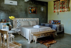 Un dormitorio con cama, un banco de madera, una mesa con mantel, una silla, una planta en maceta y un aire acondicionado de pared.