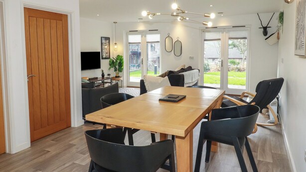 A modern dining area with a wooden table and black chairs, a ceiling light fixture, and a view of the outdoors through glass doors. - Treveglos (Truro)