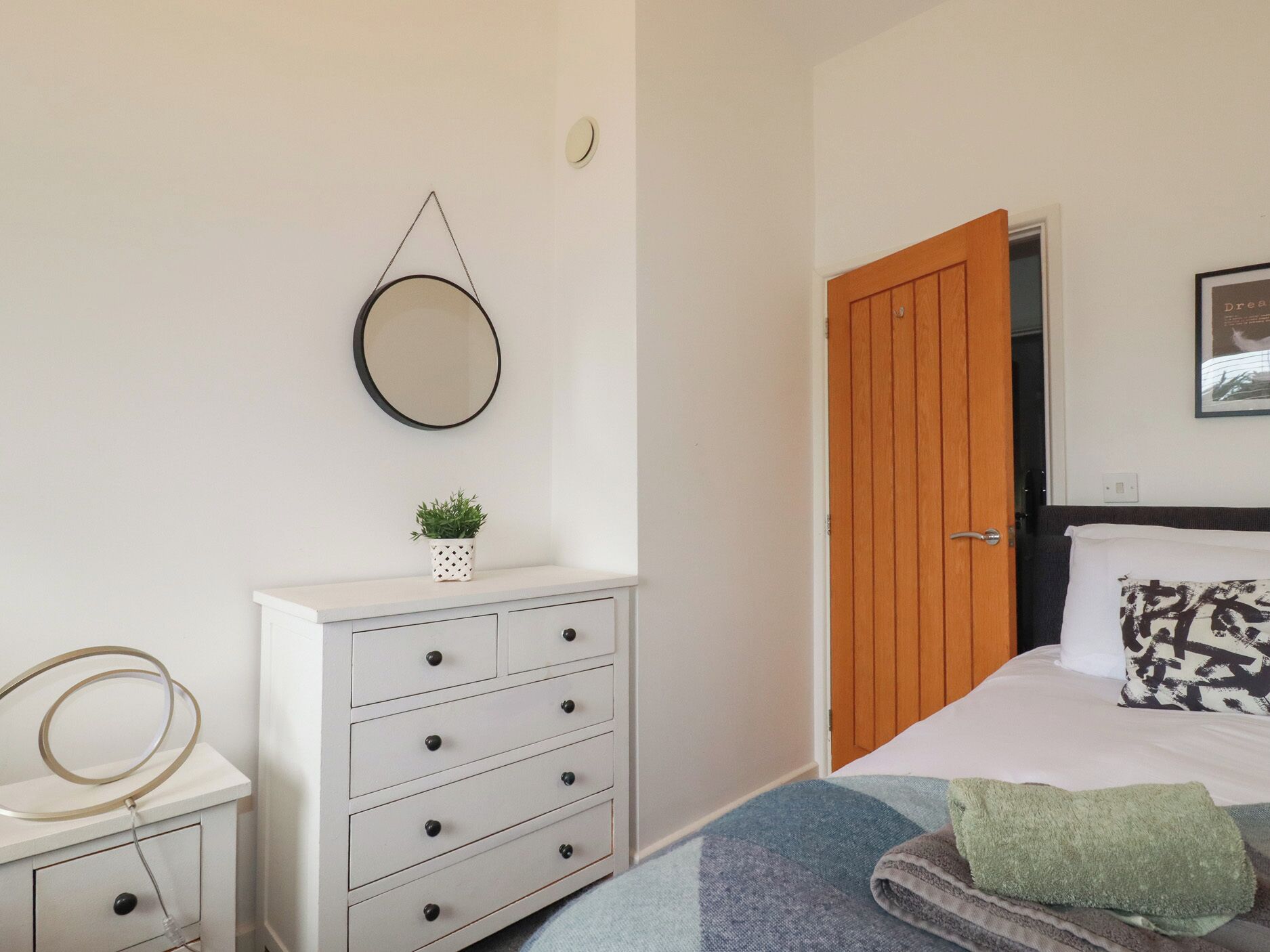 A bedroom with a bed, a dresser with drawers, a round mirror, a small potted plant, and a wooden door.