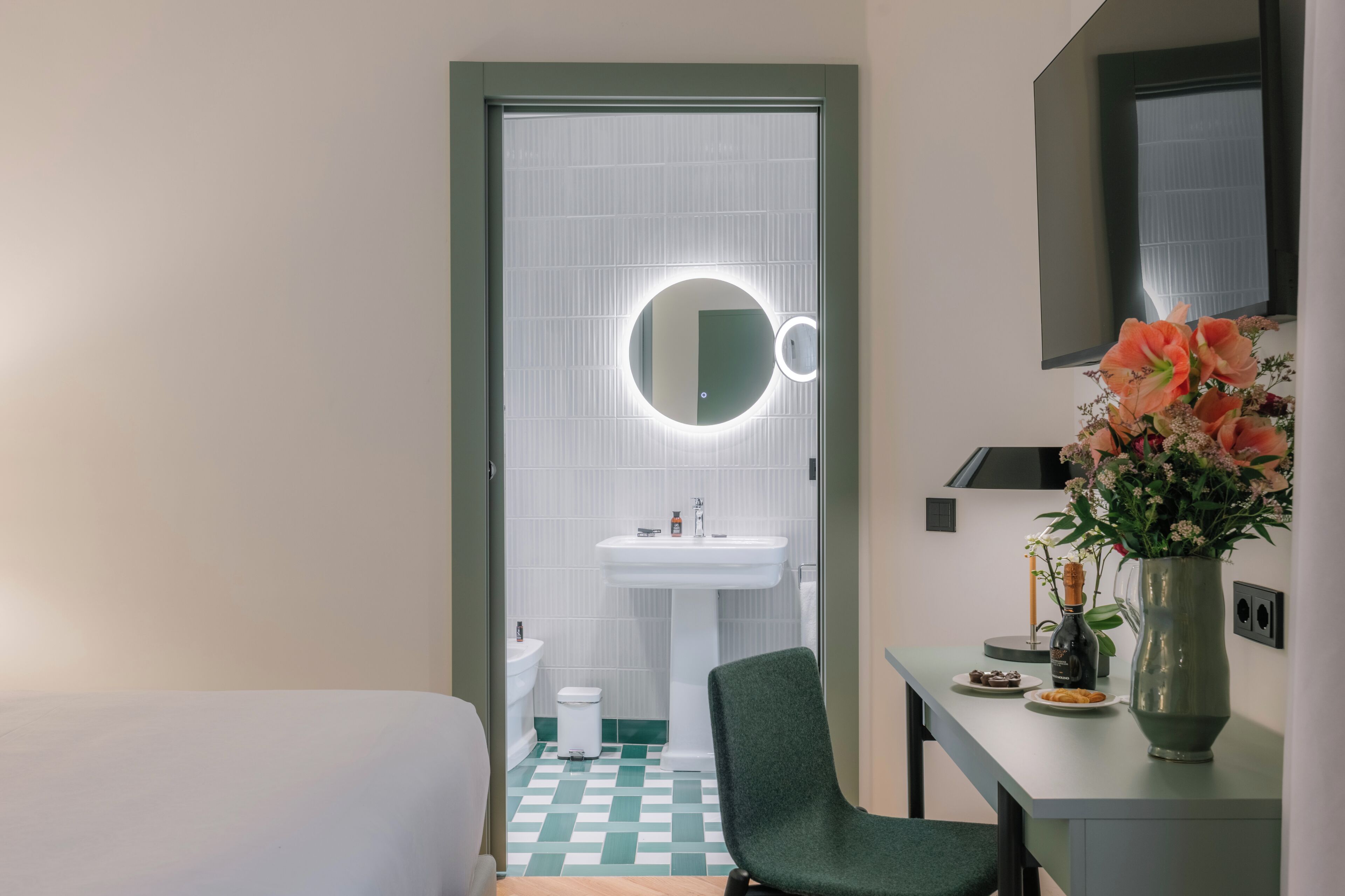 A hotel room with a bed, a green chair, a small table with a vase of flowers, a mirror, and a sink.