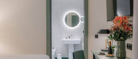 A hotel room with a bed, a green chair, a small table with a vase of flowers, a mirror, and a sink.