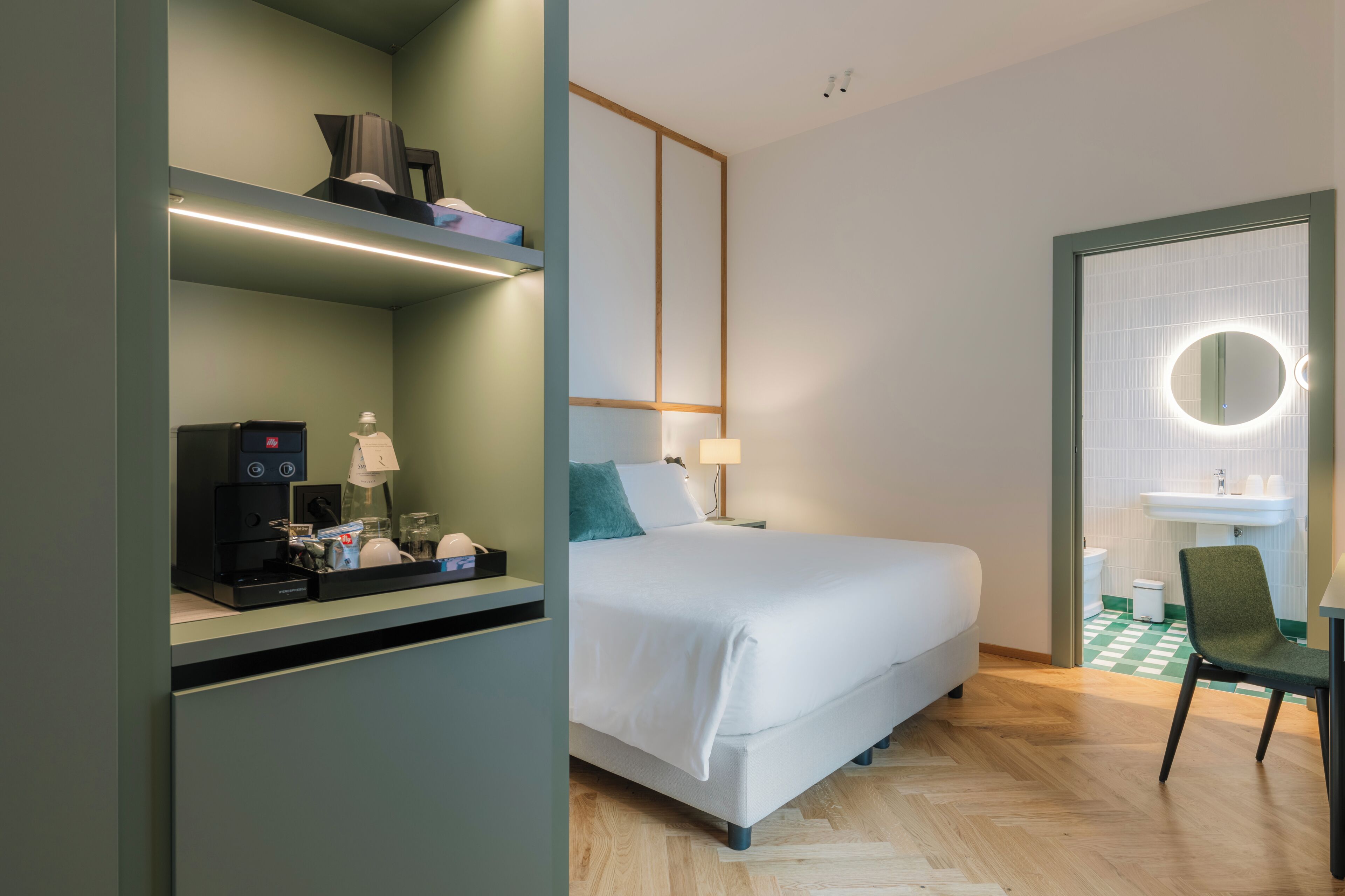 A hotel room with a bed, a desk with a coffee machine, a mirror, and a bathroom with a sink and toilet.