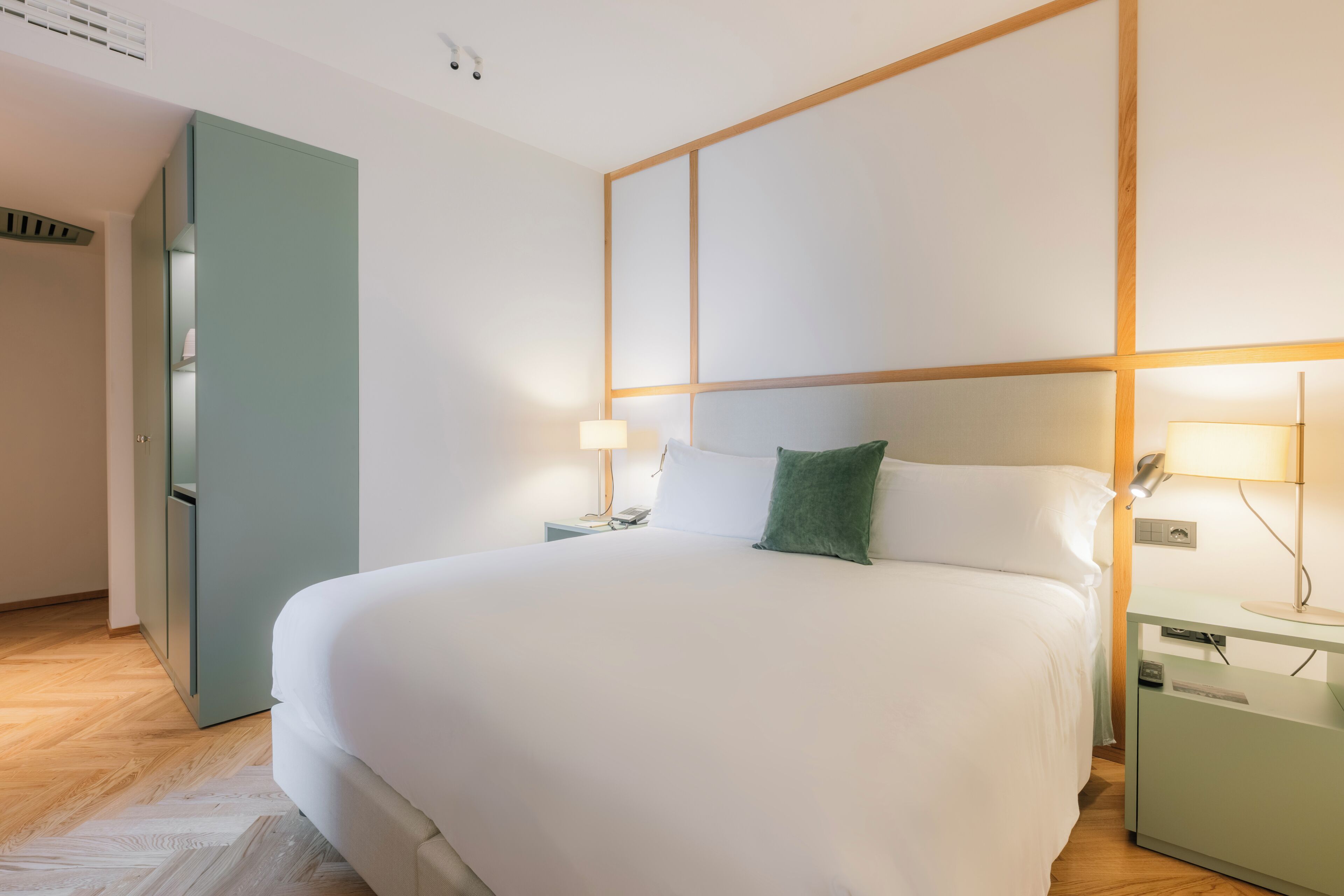 Un lit bien fait, avec du linge de lit blanc, deux lampes de chevet, un oreiller vert et une petite table de chevet avec une lampe.