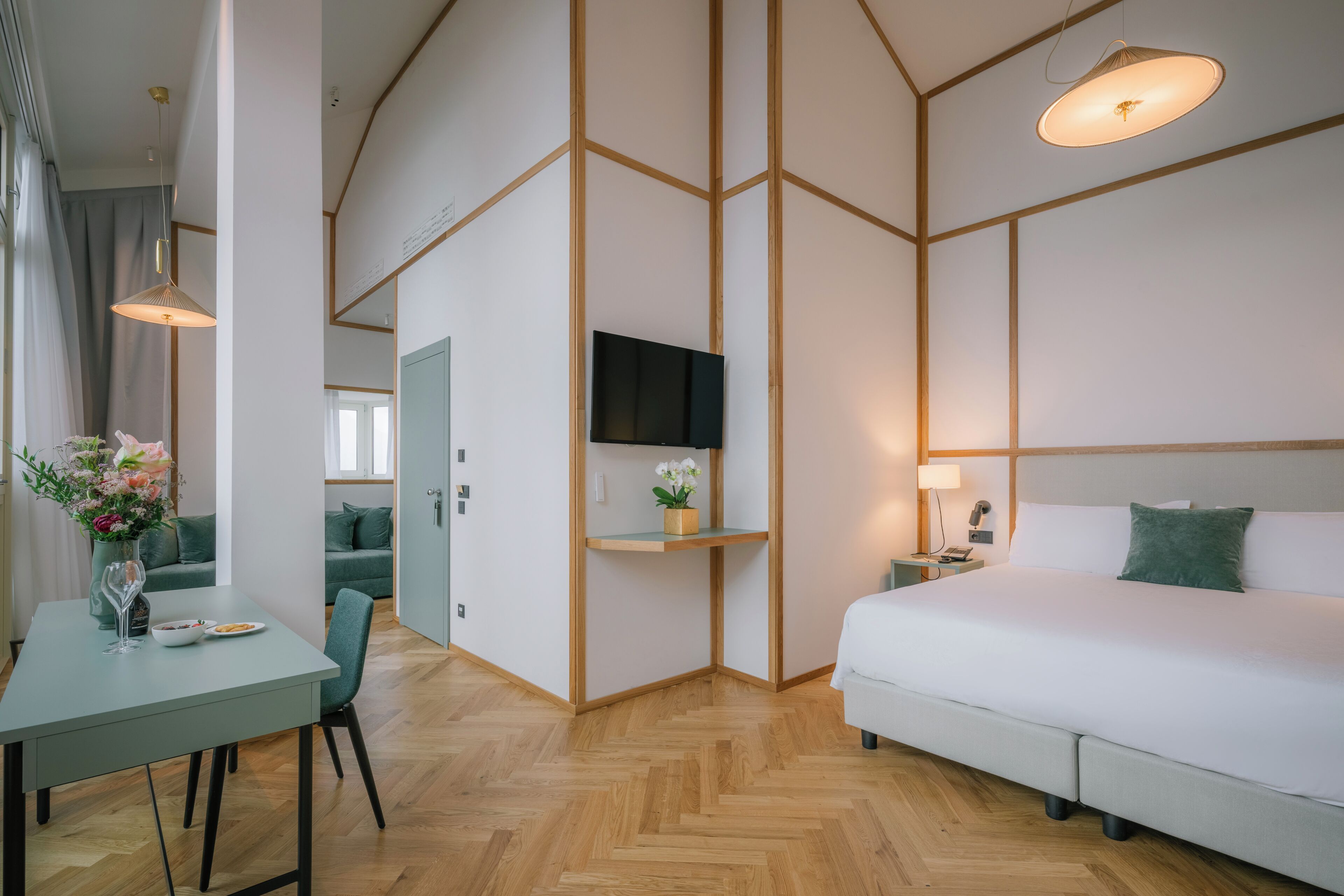 Une chambre d’hôtel moderne dotée d’un grand lit, d’un téléviseur fixé au mur, d’un coin repas avec une table et des chaises, et d’un couloir menant à une autre chambre.