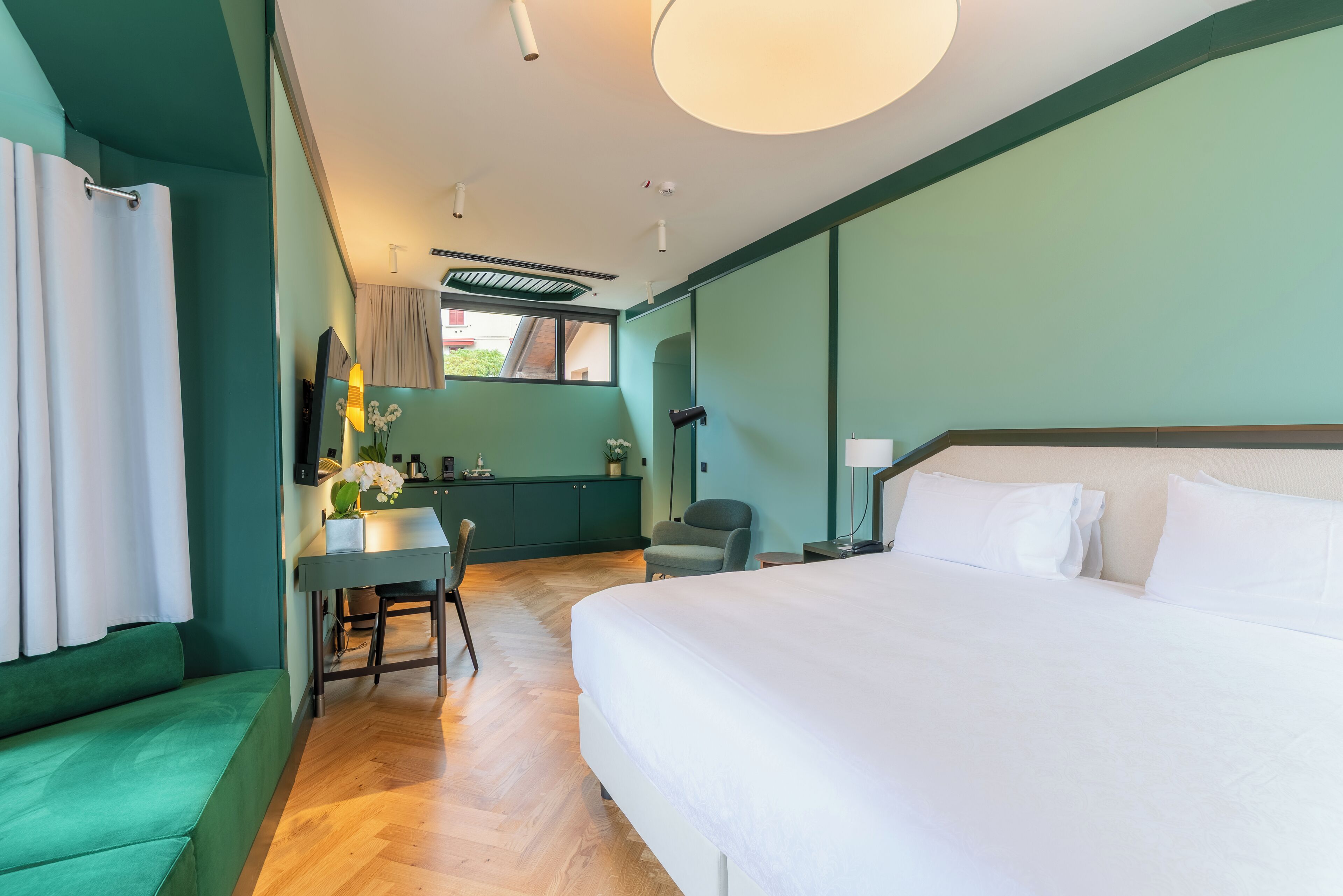 Ein Hotelzimmer mit einem großen Bett, einem Schreibtisch mit Stuhl, einem kleinen Tisch mit einer Vase und einem Fenster mit Vorhängen.