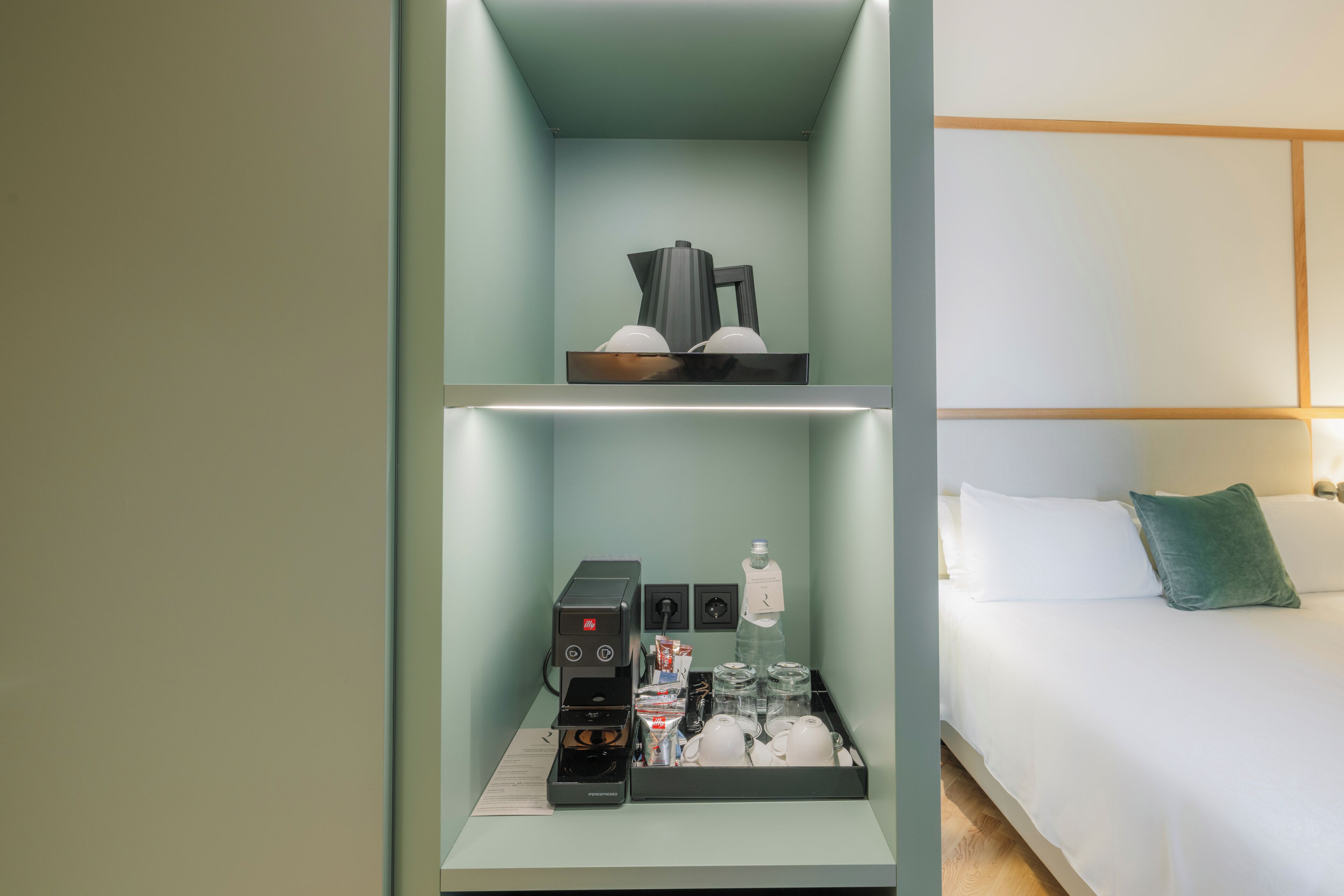 A hotel room with a bed, a nightstand with a coffee machine, and a shelf with various items.