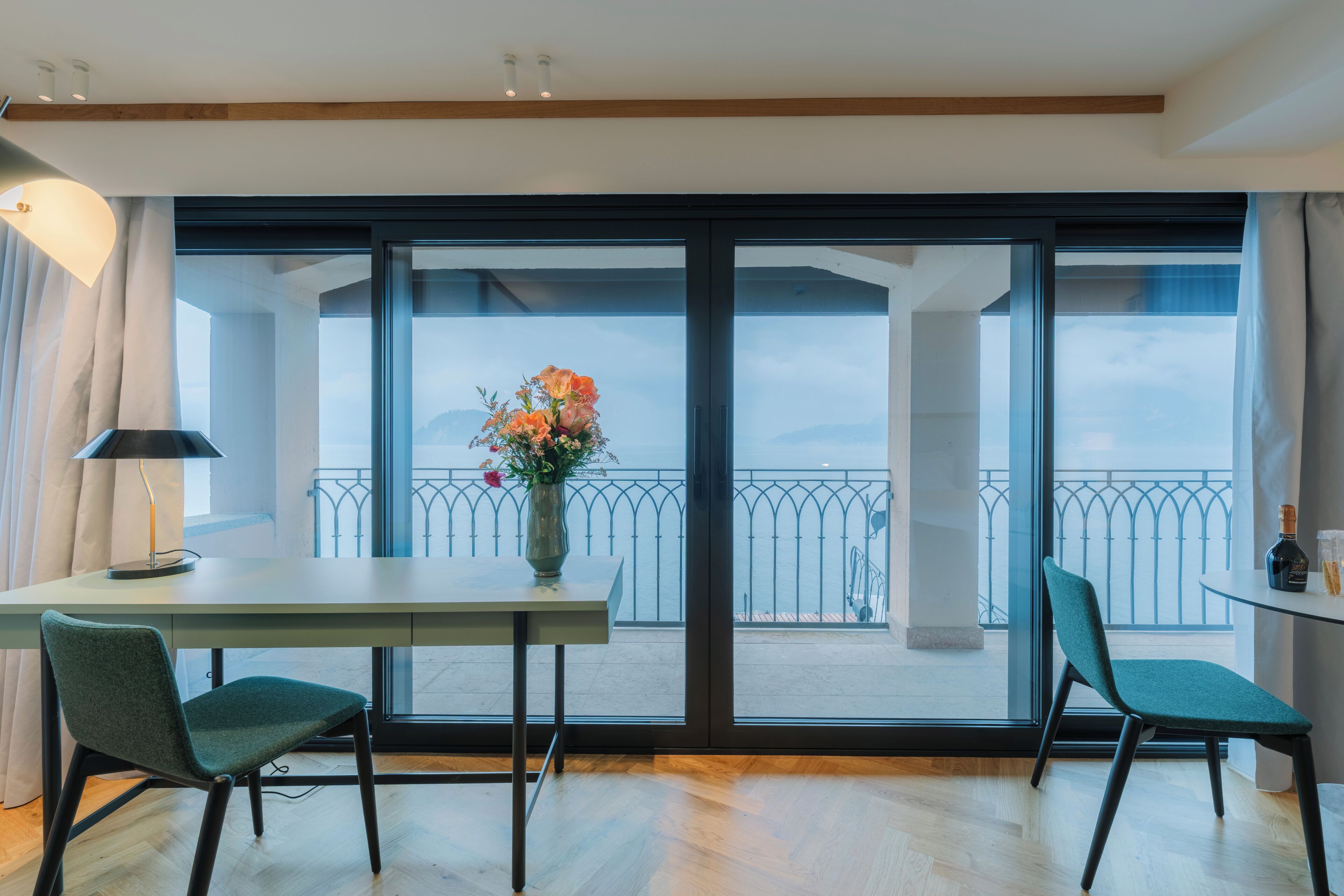 Een kamer met balkon, een tafel met stoelen, een vaas met bloemen en een lamp.
