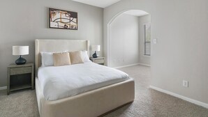 A bedroom with a bed, two bedside tables, a nightstand, a painting on the wall, and an arched doorway.