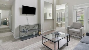 A modern living room with a flat-screen TV, a glass coffee table, a grey sofa, and a glass shelving unit.