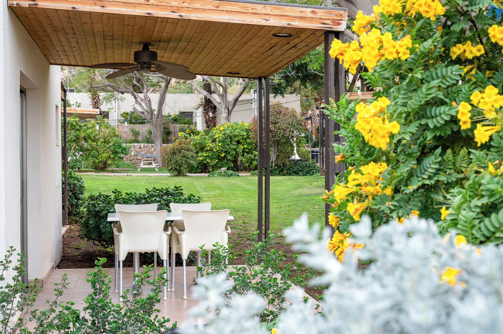 Un patio techado con mesa y sillas, un ventilador de techo y vistas a un jardín cuidado con diversas plantas y césped.