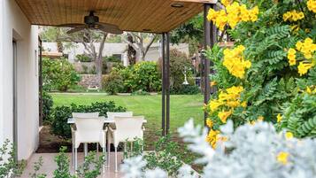 Un patio techado con mesa y sillas, un ventilador de techo y vistas a un jardín cuidado con diversas plantas y césped.