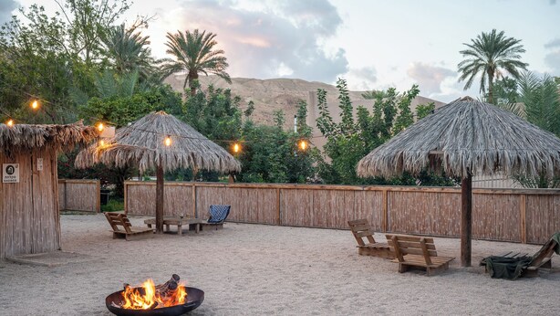 A sandy outdoor area with thatched umbrellas, wooden benches, and a fire pit. - Kalya Kibbutz Hotel (Kalya)