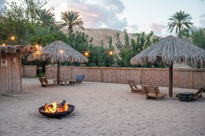 A sandy outdoor area with thatched umbrellas, wooden benches, and a fire pit.