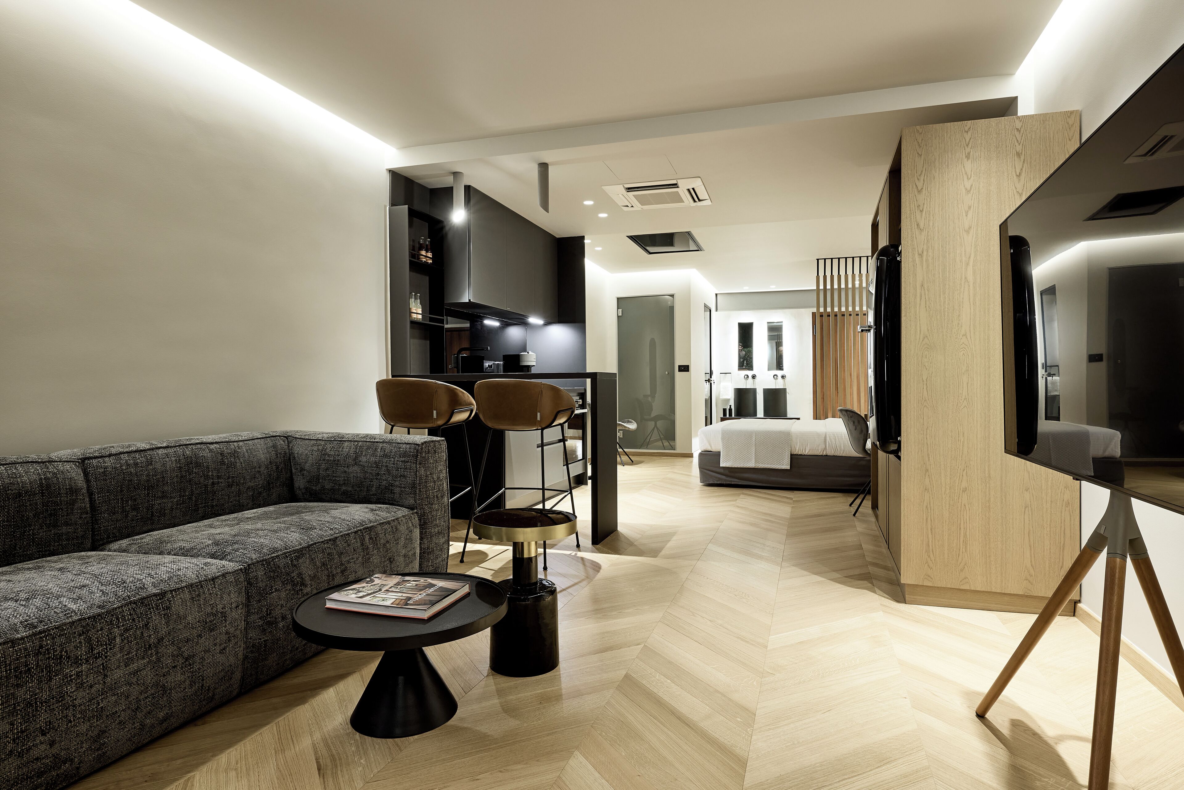 Una sala moderna con un sofá gris, una mesa de centro redonda y un televisor sobre un mueble de madera. Se aprecia una zona de cocina con encimera negra y sillas. En el fondo se ve una cama con edredón blanco.