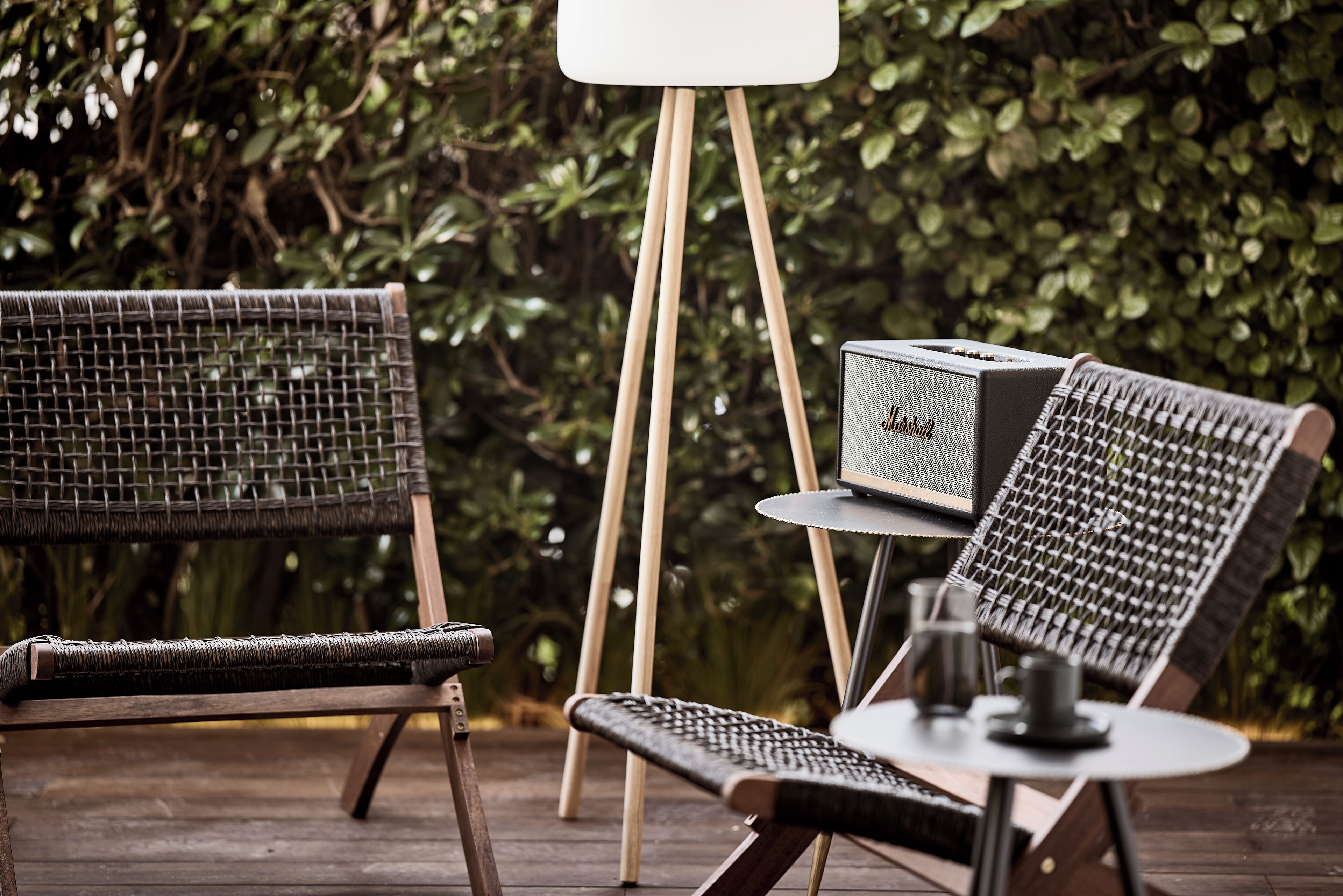 A wicker chair and a table with a vintage radio on a wooden deck surrounded by greenery.