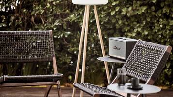 A wicker chair and a table with a vintage radio on a wooden deck surrounded by greenery.