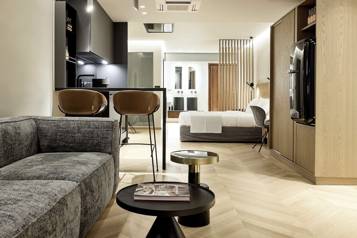 A modern hotel room with a sofa, two chairs, a small table with a book, a bed, a wardrobe, and a kitchenette.