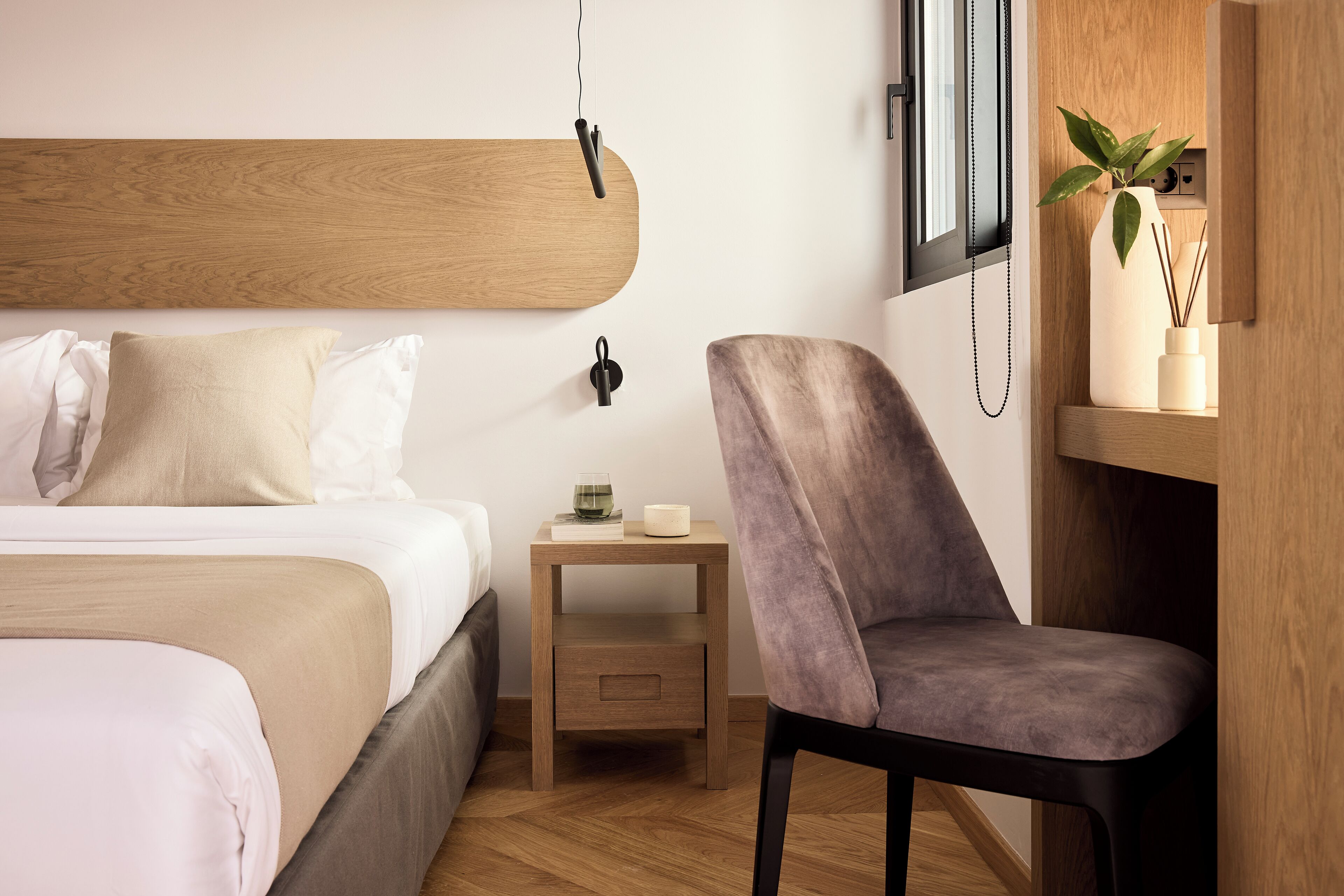 Een moderne hotelkamer met een houten hoofdeinde, een nachtkastje met een lamp, een stoel en een kleine plant op een plank.