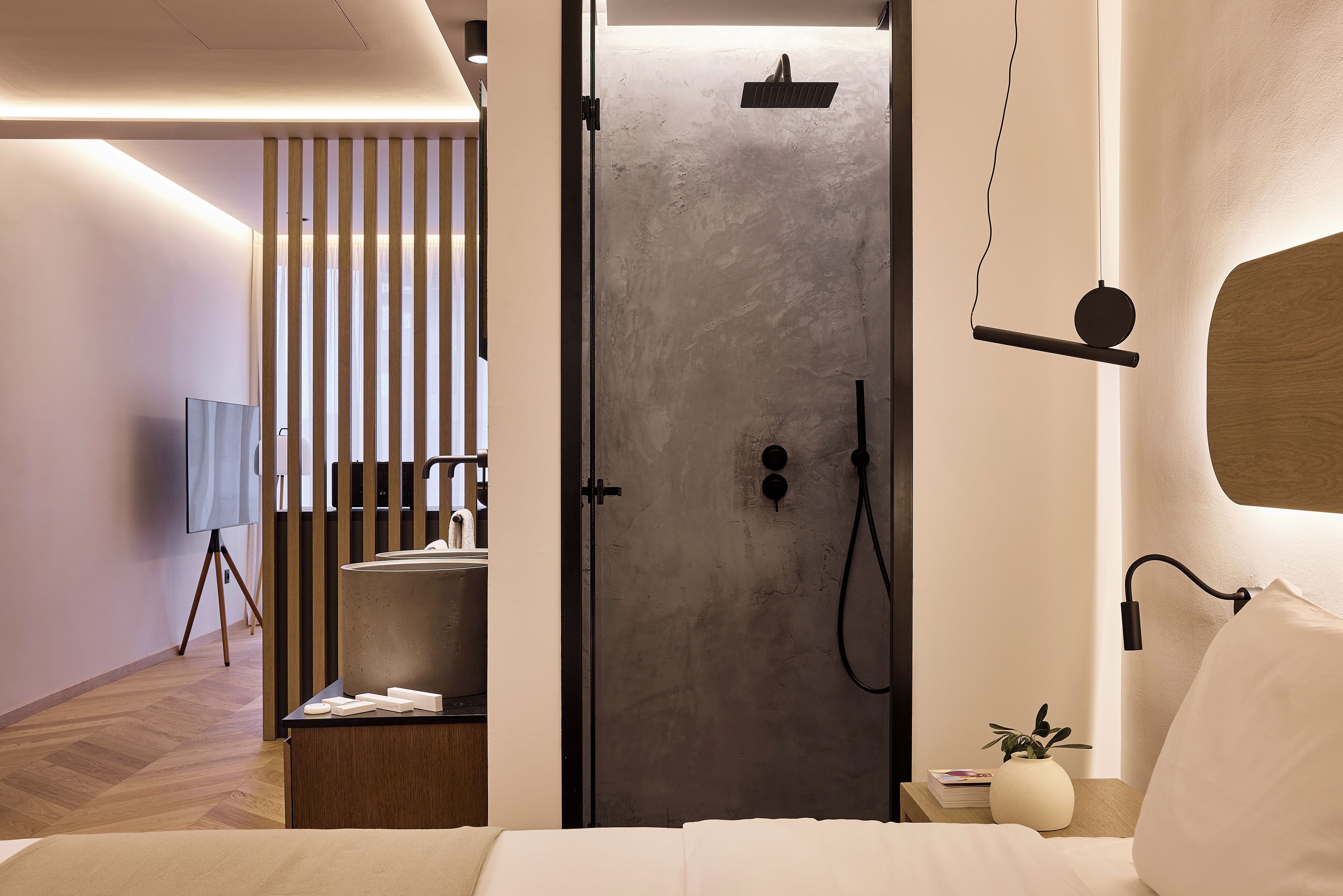 A modern hotel room with a minimalist design, featuring a large bed, a bedside table with a lamp, a small plant, and a shower area with a towel rack.