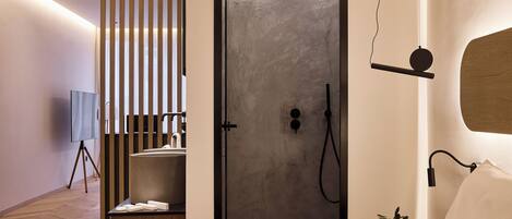 A modern hotel room with a minimalist design, featuring a large bed, a bedside table with a lamp, a small plant, and a shower area with a towel rack.