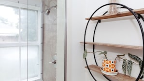A bathroom with a glass shower door, a white tiled wall, and a wooden shelf with a potted plant.