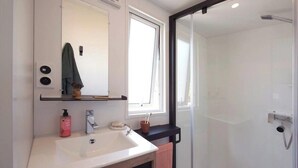 Un baño moderno con lavamanos blanco, un espejo, una ventana y ducha con mampara de vidrio.