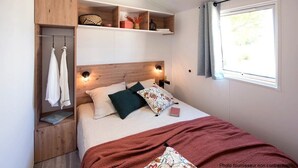 Un dormitorio ordenado con cabecera de madera, mesita de noche con lámpara y ventana con vista a vegetación.