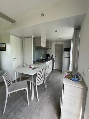 A compact kitchen with a dining area, featuring a white table, chairs, and a small island with a microwave and kettle. - ZEcamping 3 étoiles familial piscine couverte locatif LV BBQ 29m2 (Mont-prés-Chambord)