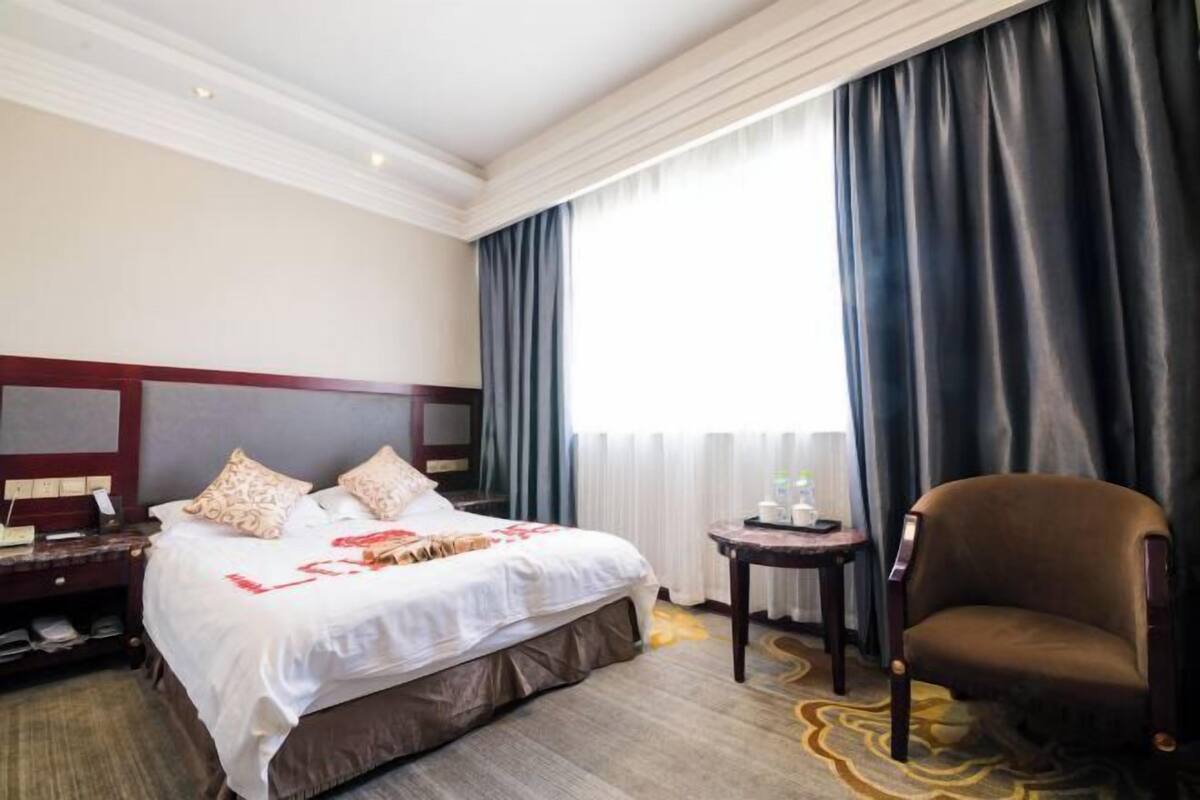 Une chambre d’hôtel avec un grand lit, une chaise, une petite table et une fenêtre avec des rideaux.