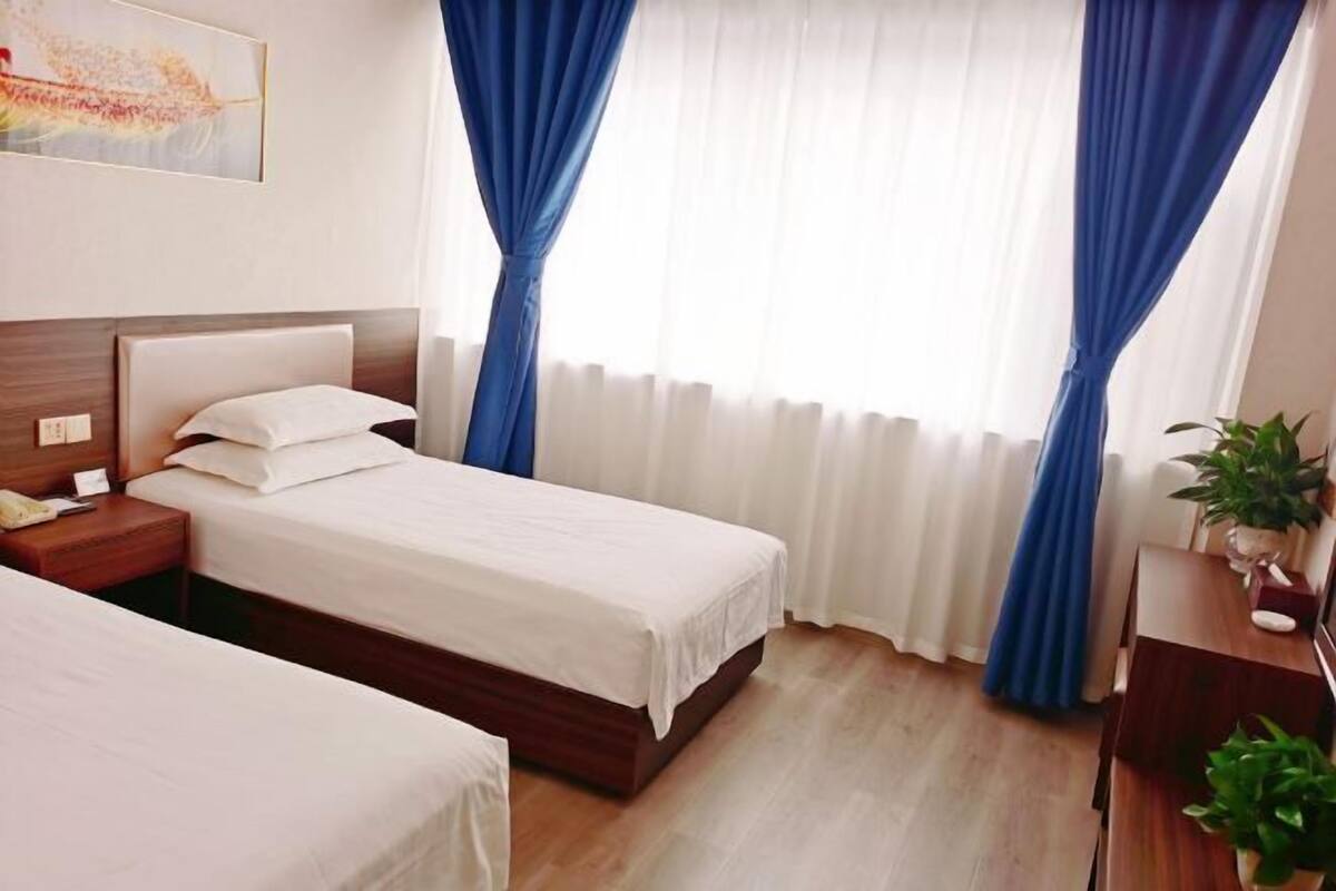 Une chambre d’hôtel avec deux lits, une tête de lit en bois, une table de chevet avec une lampe, une fenêtre avec des rideaux bleus et un tableau au mur.
