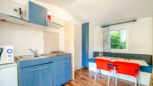 Une cuisine compacte équipée d’un évier, d’une cuisinière et de meubles de rangement bleus. On y trouve également un coin repas avec une table et deux chaises rouges. Une fenêtre avec des rideaux est visible.
