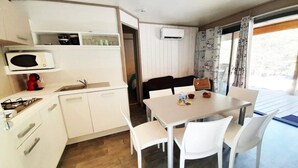 Cozinha compacta com micro-ondas, fogão e armários brancos. Há uma área de refeições com mesa e cadeiras, e uma janela com cortinas.