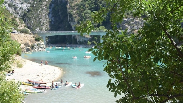 A beach with kayaks and people, a bridge, and a mountainous backdrop.